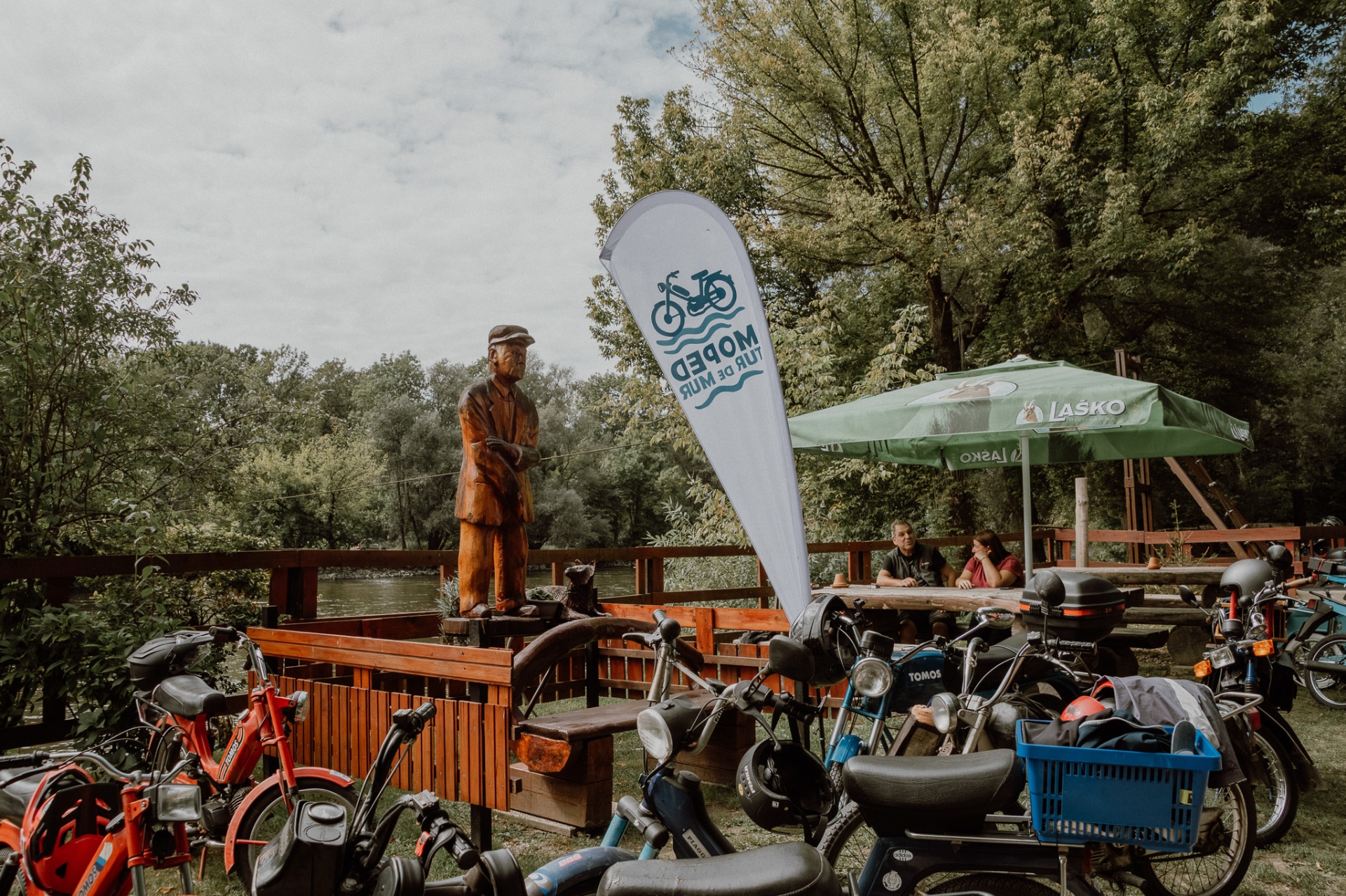 Zastava MOPED Mur ob leseni skulpturi ribiča na bregu Mure