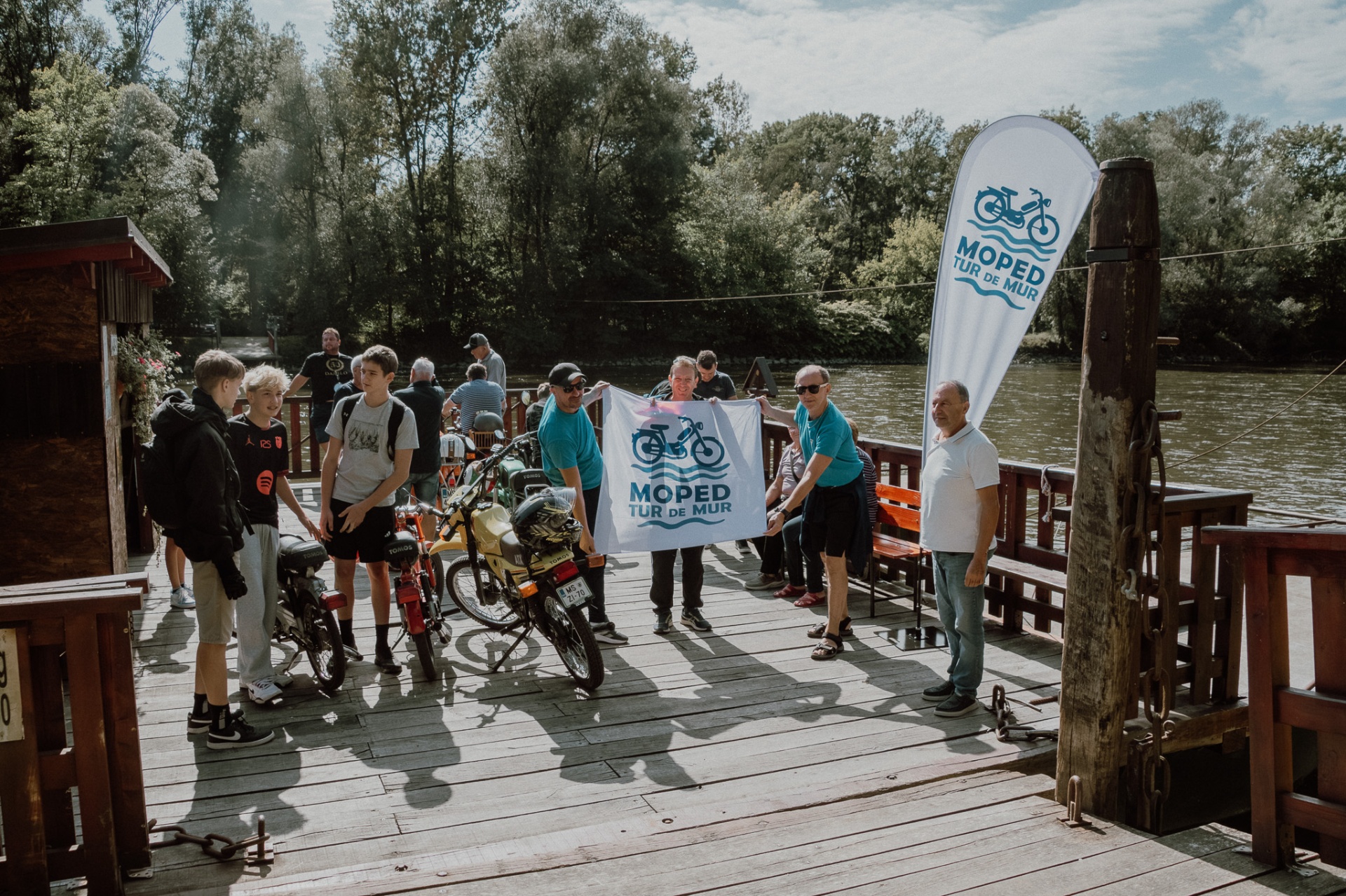 Udeleženci držijo zastavo MOPED Mur na pomolu broda